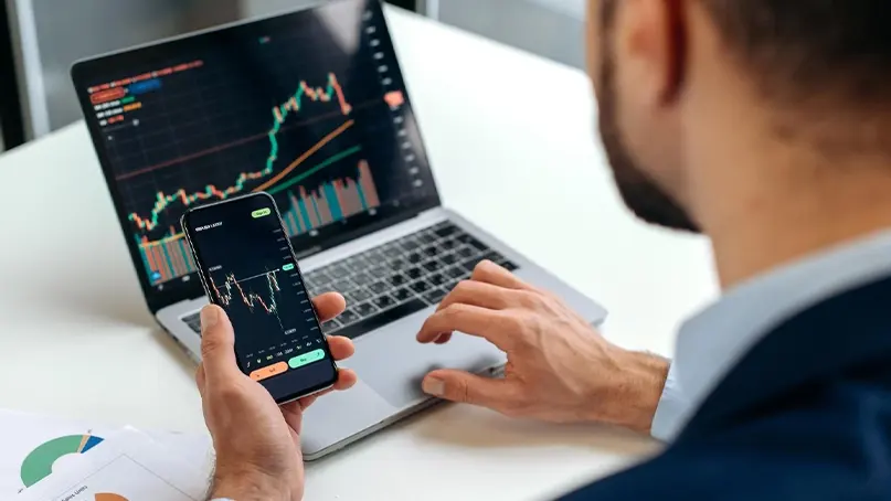 A person checking price charts on a smartphone with a laptop showing more charts beside it